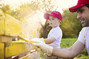 Cute little boy and his father in red caps painting wooden fence together on sunny day in nature