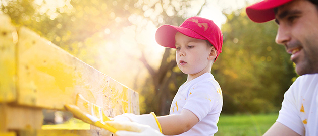 Cute little boy and his father in red caps painting wooden fence together on sunny day in nature