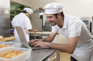 chef in white hat standing in kitchen and typing on a laptop computer