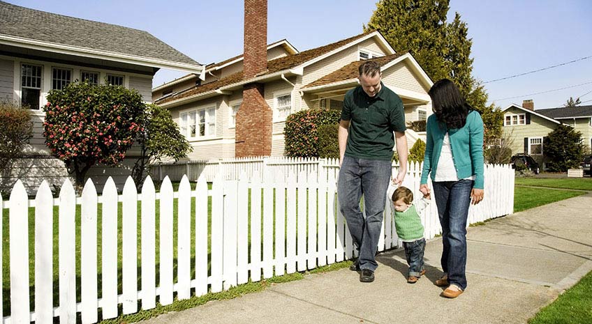 young family of dad, mom and toddler walk on the sidewalk by a white picket fence
