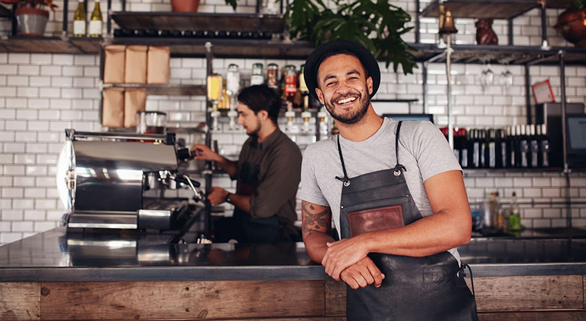 Coffee shop worker smiling to camera, standing at the counter. Happy young man in apron and hat leaning to cafe counter, with waiter working in background.