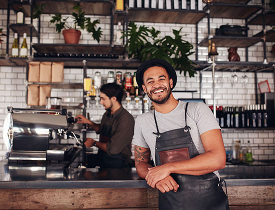 Coffee shop worker smiling to camera, standing at the counter. Happy young man in apron and hat leaning to cafe counter, with waiter working in background.