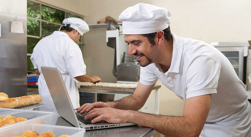 chef in white hat standing in kitchen and typing on a laptop computer