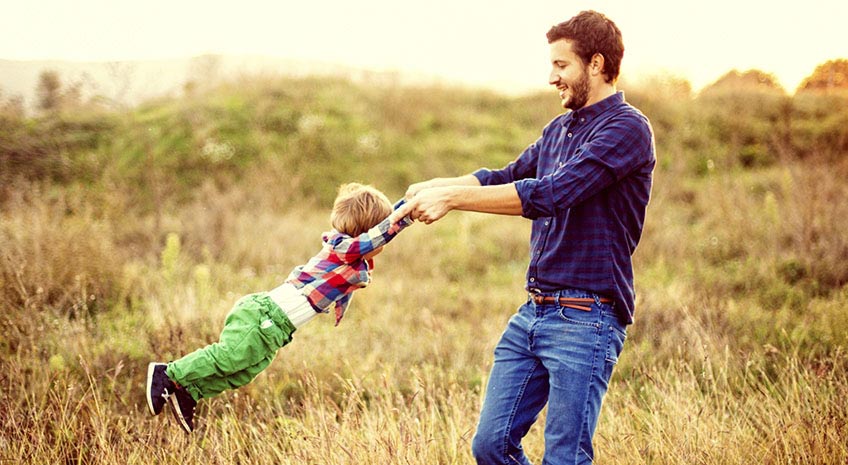 father swings son around in a circle by holding his arms while standing in a grassy field