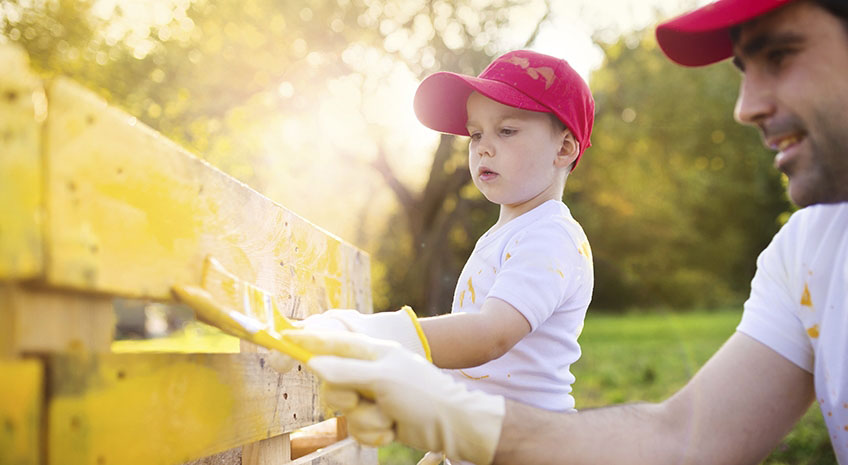 Cute little boy and his father in red caps painting wooden fence together on sunny day in nature