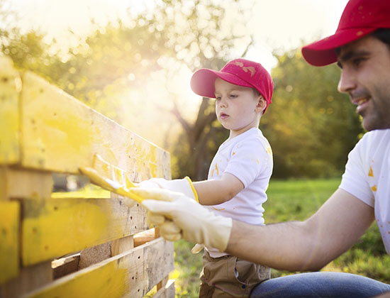 Cute little boy and his father in red caps painting wooden fence together on sunny day in nature