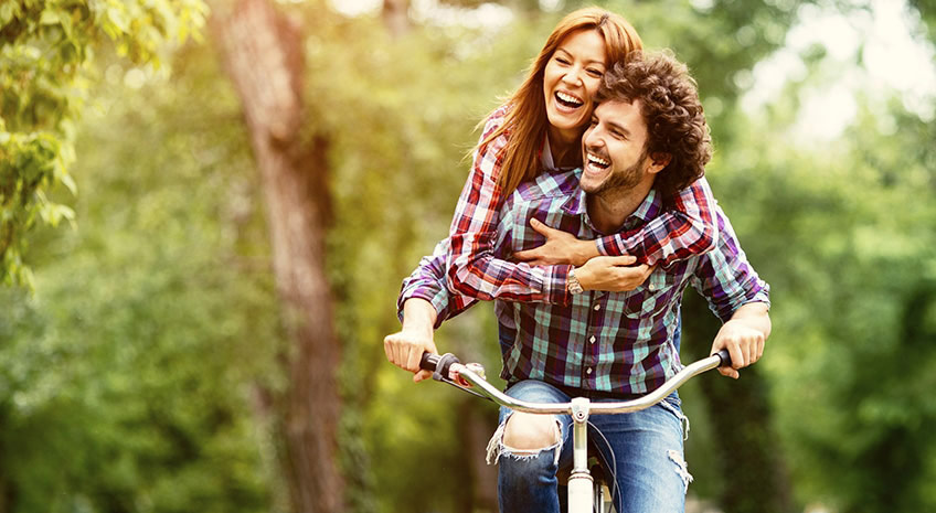 smiling and laughing man and woman riding together on bicycle through sunny wooded area