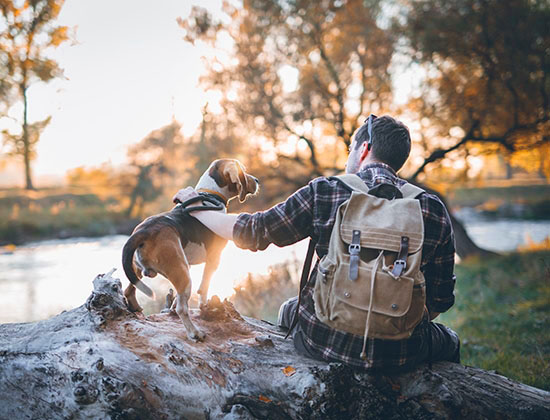 Man hiker and his dog taking a rest from walking, sitting by the river, enjoying the view and sunset