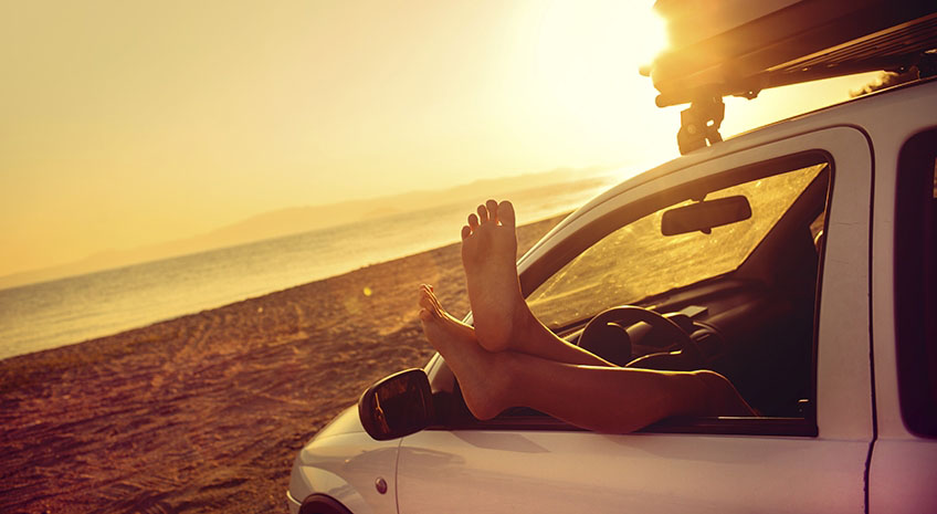 feet hanging out the drivers window of a car while loverlooking a beach sunset
