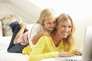 smiling woman in yellow shirt laying on floor and looking at computer with daughter lying on her back