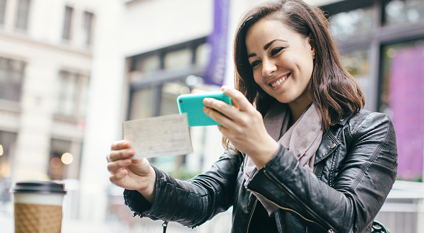 A beautiful Caucasian adult woman sits in a New York city park, taking a picture of a check with her smart phone for a Remote Deposit Capture. She smiles, wearing modern stylish clothing with darker and black colors.