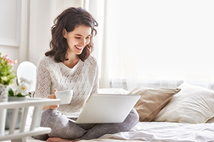 Happy casual beautiful woman working on a laptop sitting on the bed in the house.