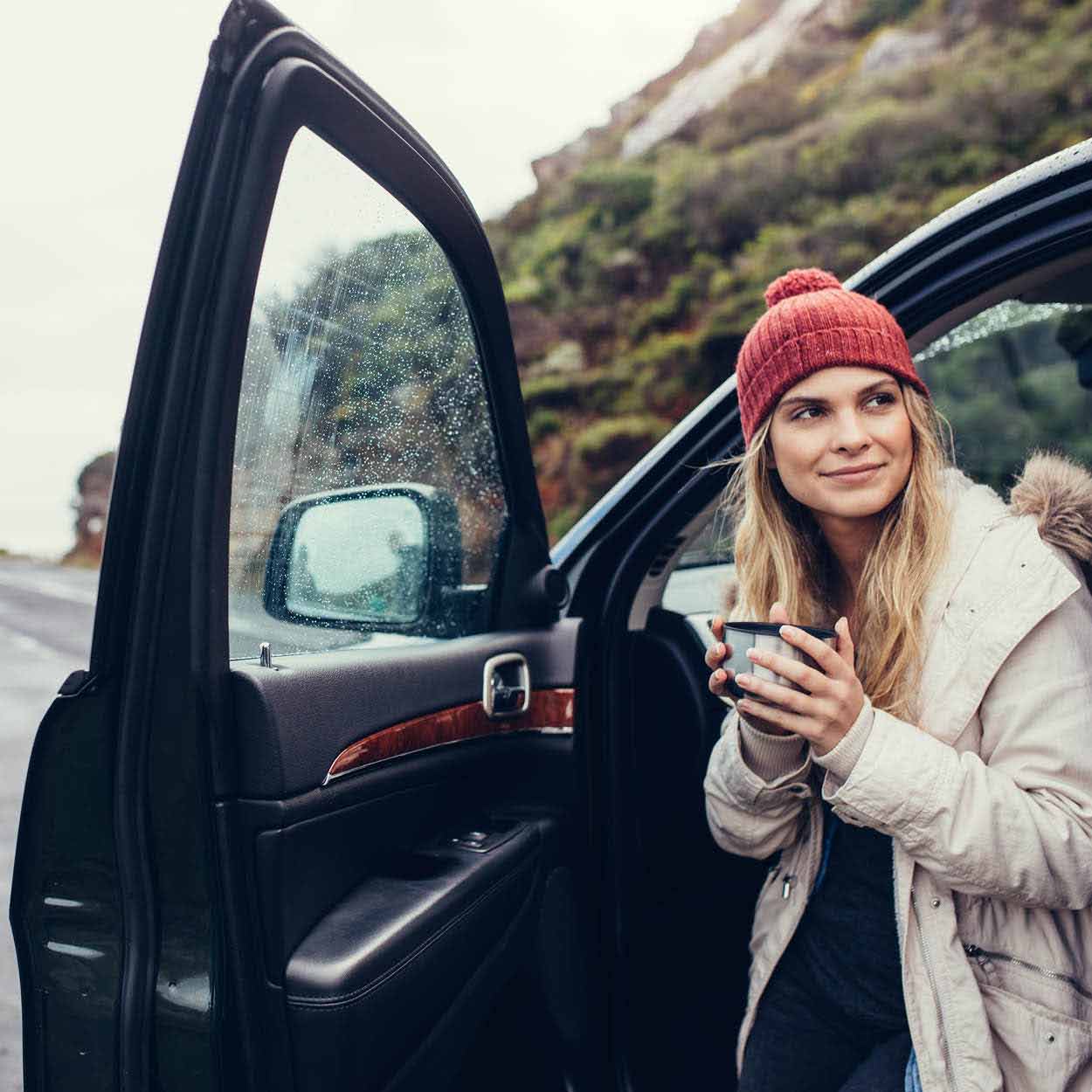 woman holding coffee in hat by car