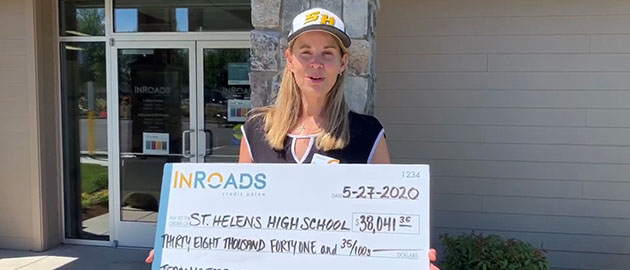 Middle aged woman with blonde hair wearing a baseball cap and holding an oversize check made out to St. Helens High School