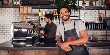 Coffee shop worker smiling to camera, standing at the counter. Happy young man in apron and hat leaning to cafe counter, with waiter working in background.