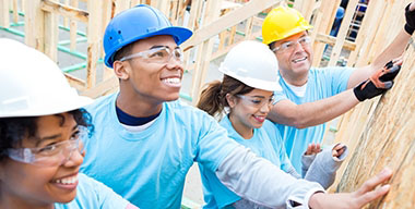 Diverse friends are volunteers working together to build home for charity. They are confidently putting up a wall in the home. They are all holding the wall. They are wearing hard hats, safety glasses and light blue volunteer t-shirts.