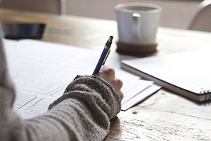 tan sweater covered arm and hand holding a blue pen and writing on papers