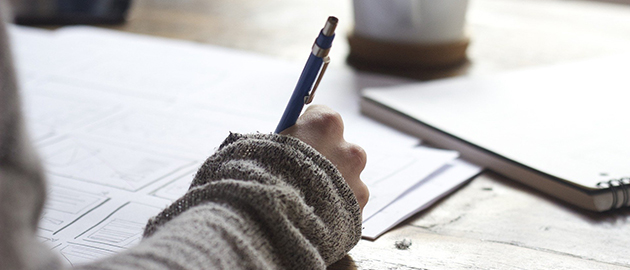 tan sweater covered arm and hand holding a blue pen and writing on papers