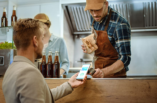 Food Truck Employee Hands Out a Freshly Made Burger to a Happy Young Male. Man in a Casual Suit is Using His Smartphone for NFC Mobile Payment Solution. Street Food Truck Selling Burgers.