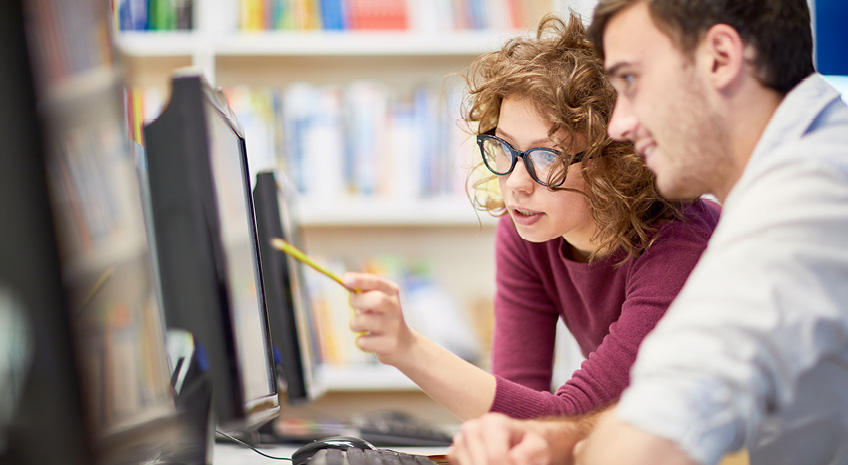 Female college student showing computer program to her classmate during a lesson at computer class at college