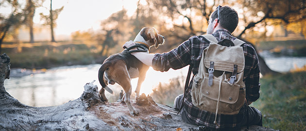 Man hiker and his dog taking a rest from walking, sitting by the river, enjoying the view and sunset