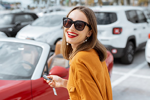 Portrait of a beautiful young woman standing with keys near the red cabriolet at the car parking outdoors. Concept of a happy car buying or renting