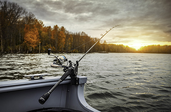 side of boat with fishing pole and line in the river with a backdrop of fall trees and sunrise under cloudy skies