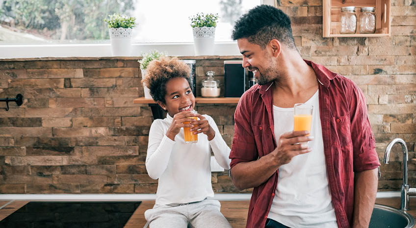 African American father and daughter sitting in the kitchen and drinking orange juice
