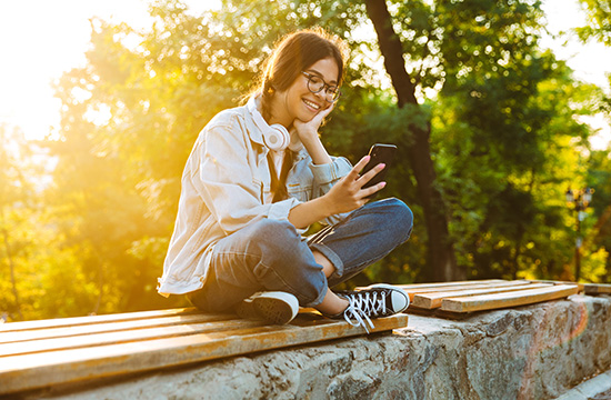 Photo of a cheerful happy cute young student girl wearing eyeglasses sitting outdoors in nature park using mobile phone.