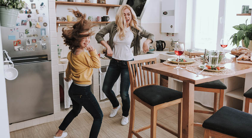 Full length portrait of charming blond woman spending time with her adorable kid. They dancing and laughing in kitchen