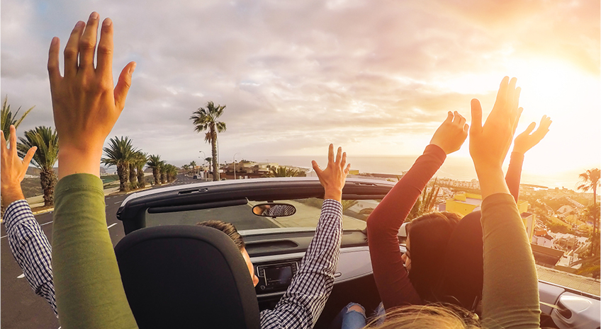 friends driving in a convertible with hands raised, in a troical place next to water and palm trees