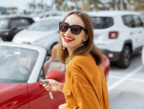 brunette woman with red lipstick and glasses, holds keys to a red convertible in a car lot