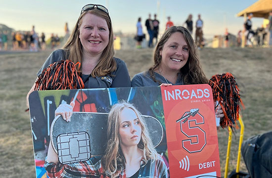 two female employees standing outside a football field holding pom poms and a large debit card with the school logo