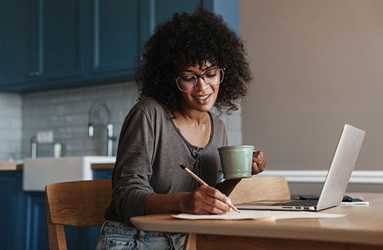 Smiling young female entrepreneur working on laptop at the kitchen table, drinking coffee