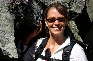 middle aged female staff member in white sweatshirt, backpack and sunglasses standing next to rock