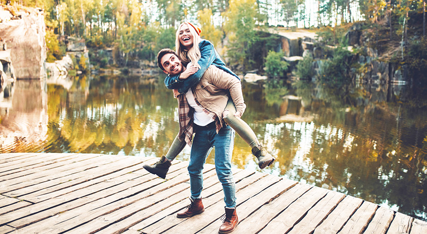 man in brown jacket giving a woman in a yellow beanie a piggy back on a dock in the fall