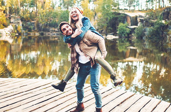 man in brown jacket giving a woman in a yellow beanie a piggy back on a dock in the fall