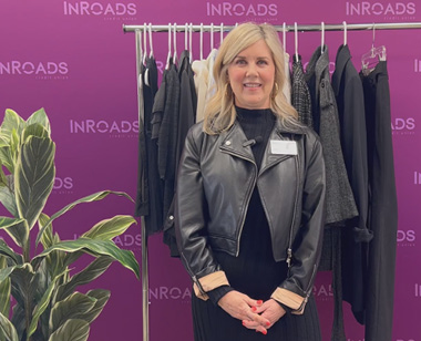 Chief Admin Officer Amy Howard stands in front of purple InRoads backdrop with a rolling rack of clothing and a green plant