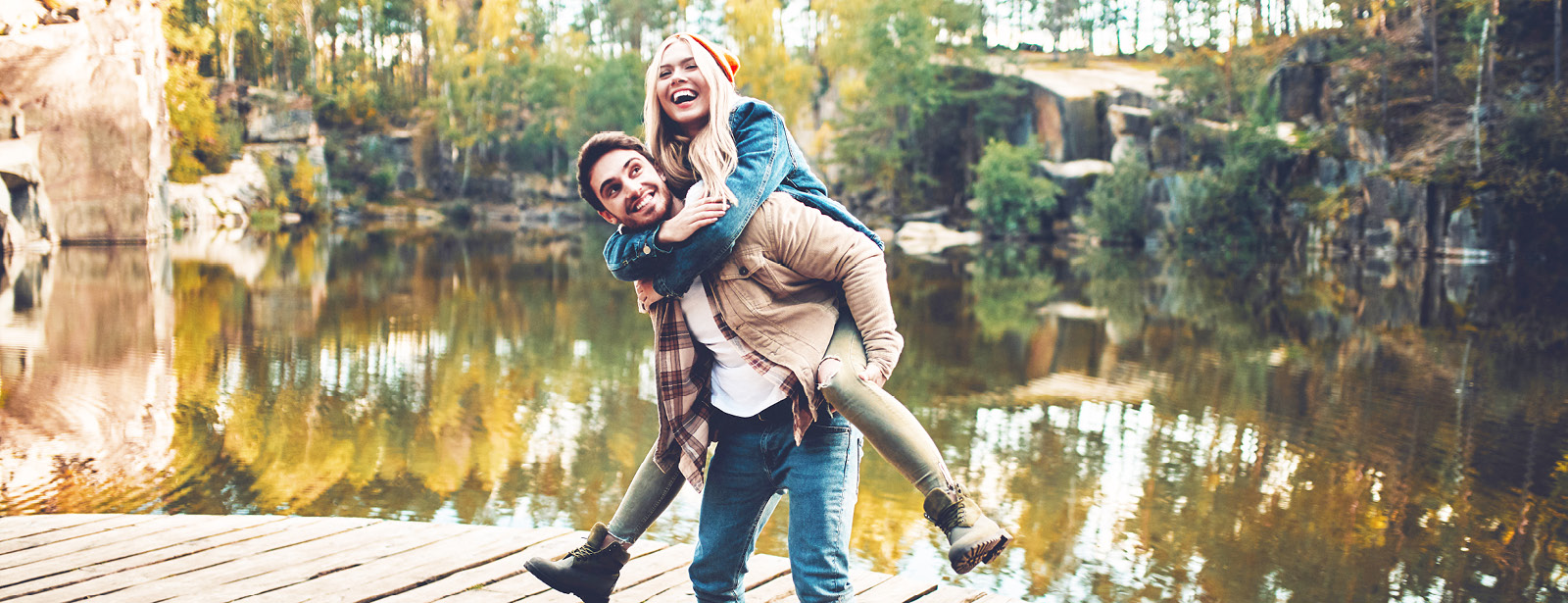 man in brown jacket giving a woman in a yellow beanie a piggy back on a dock in the fall