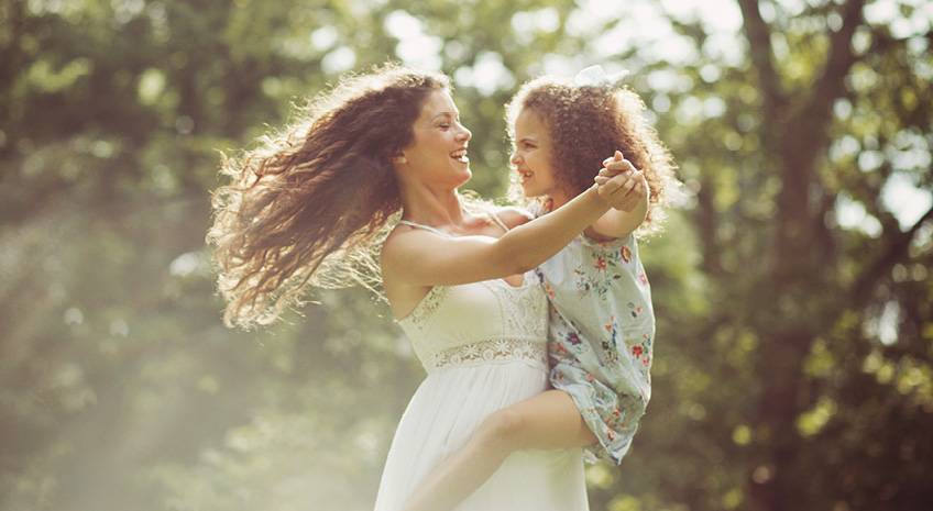 mother with long curly hair dancing at park with daughter in a dress