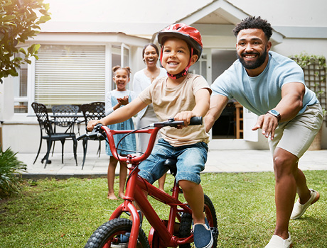 Learning, bicycle and proud dad teaching his young son to ride while wearing a helmet for safety in their family home garden. Active father helping and supporting his child while cycling outside stock photo. Family, Back Yard, Playing, Child, Happiness