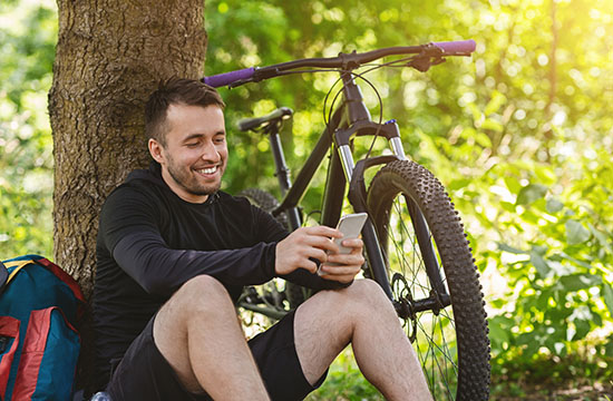 Smiling guy sitting next to bike under the tree, chatting on smartphone, copy space