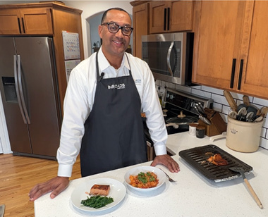 InRoads President/CEO, Nathan Cox, in a white button down and InRoads branded apron posing with cooked dishes for DIY with InRoads