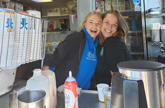 Two young adult women smiling at the camera with coffee making supplies around them