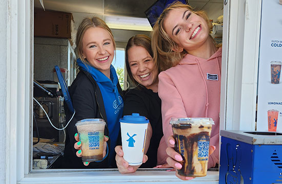 Three young adult women holding out different coffee drink through coffee shop window