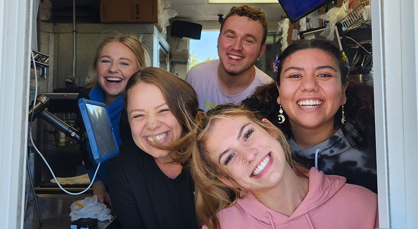 Four young women and one young man smiling at the camera through the delivery window at coffee drive thru