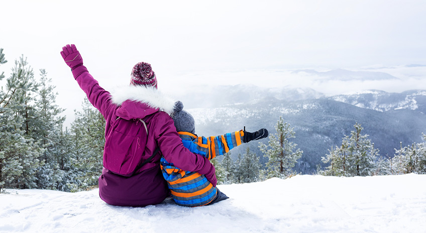 adult and child sitting on a snowy hill, happy, bundled in snow clothes to stay warm