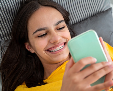 Portrait of a smiling teen girl with braces using her smartphone lying in bed