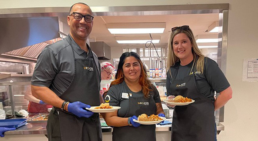 three inroads team members holding plates of food and wearing gray aprons