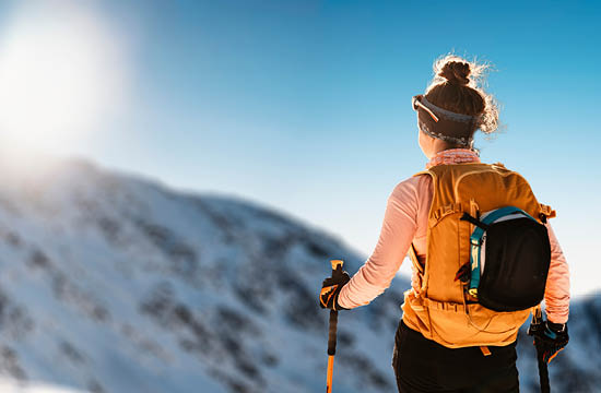 woman in yellow jacket, wearing a back pack, looks at mountains while skiing